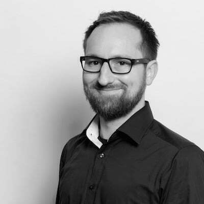 Auto-generated description: A man with glasses and a beard is smiling while wearing a dark button-up shirt in a black-and-white photo.